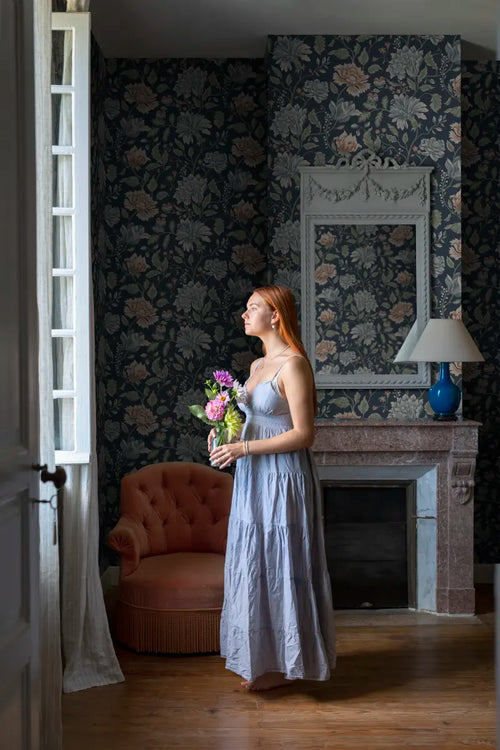 Annabelle wallpaper by Sandberg in navy floral styled in a period room with a woman, pink armchair and marble fireplace