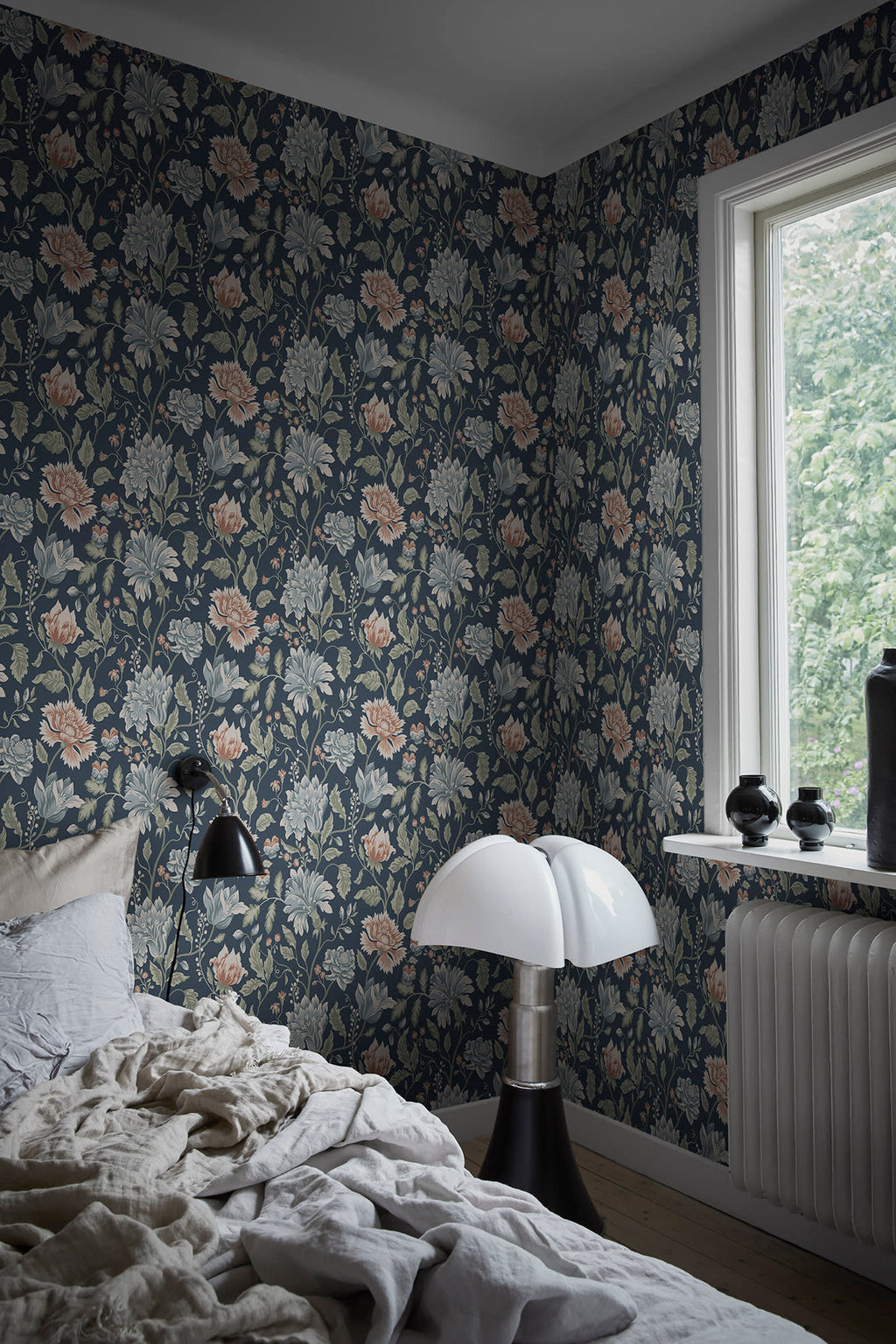 Annabelle wallpaper by Sandberg in dark floral behind a bed in a moody bedroom with a domed lamp and window