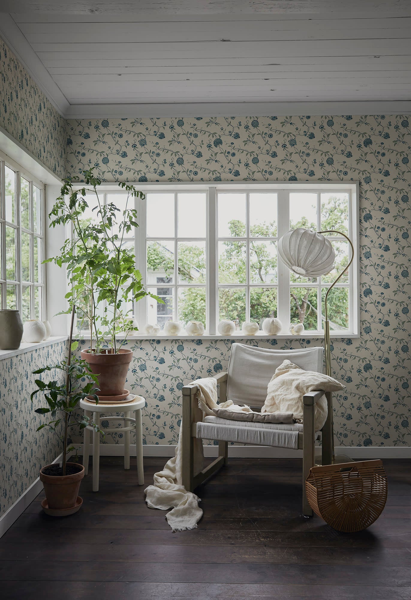 Adelaide wallpaper by Sandberg, blue floral print in a bright sunroom with a linen chair and potted plants