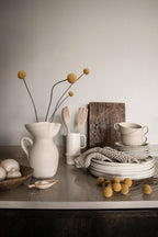 Rand Stripe wallpaper by Sandberg in warm greige, styled behind a kitchen counter with stoneware jug, bowls and dried billy buttons