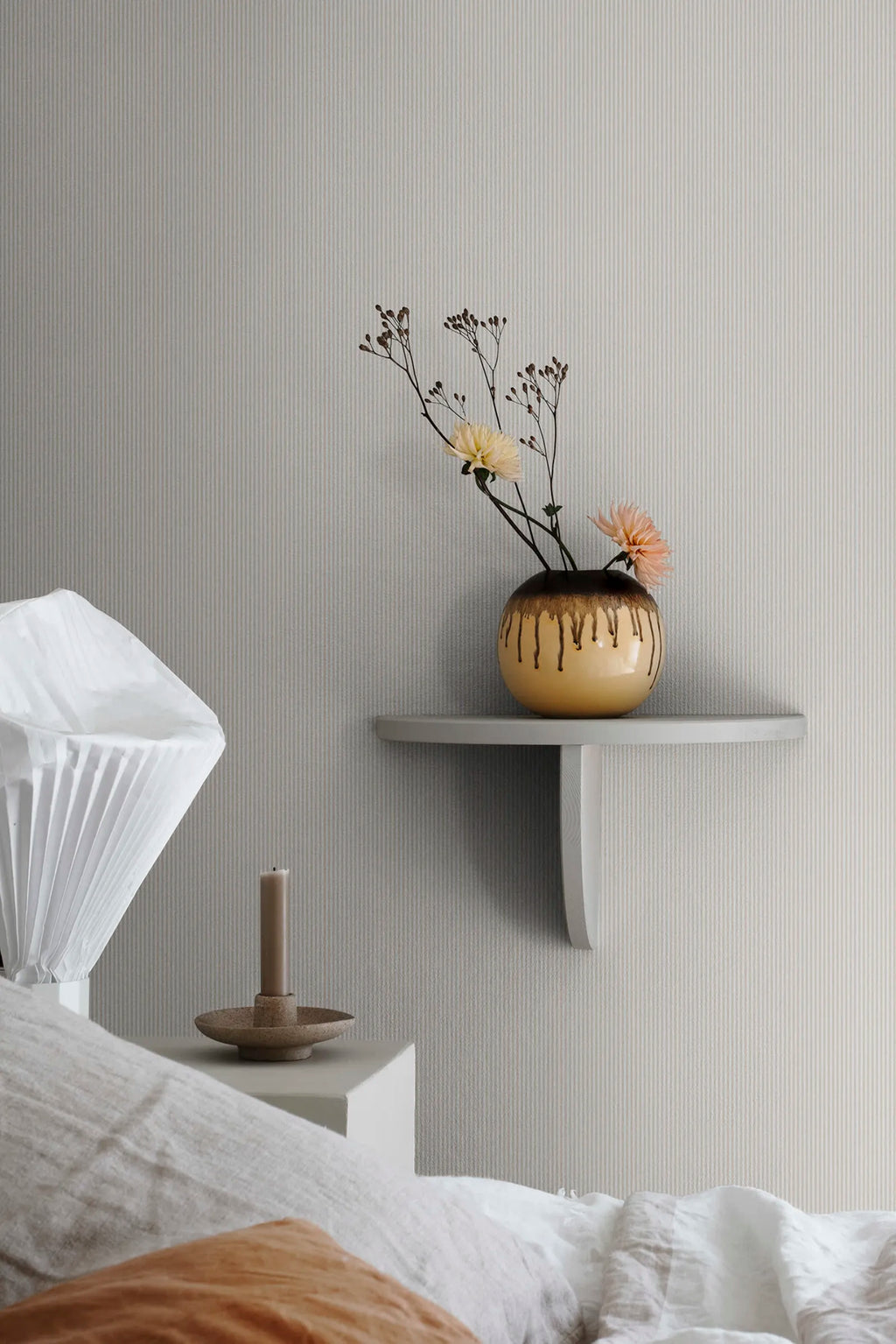 Rand Stripe wallpaper by Sandberg in soft sand, styled above a bed beside a white wall shelf with drip-glazed vase