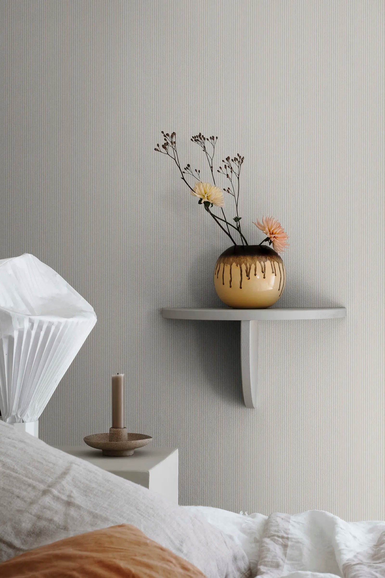 Rand Stripe wallpaper by Sandberg in soft sand, styled above a bed beside a white wall shelf with drip-glazed vase