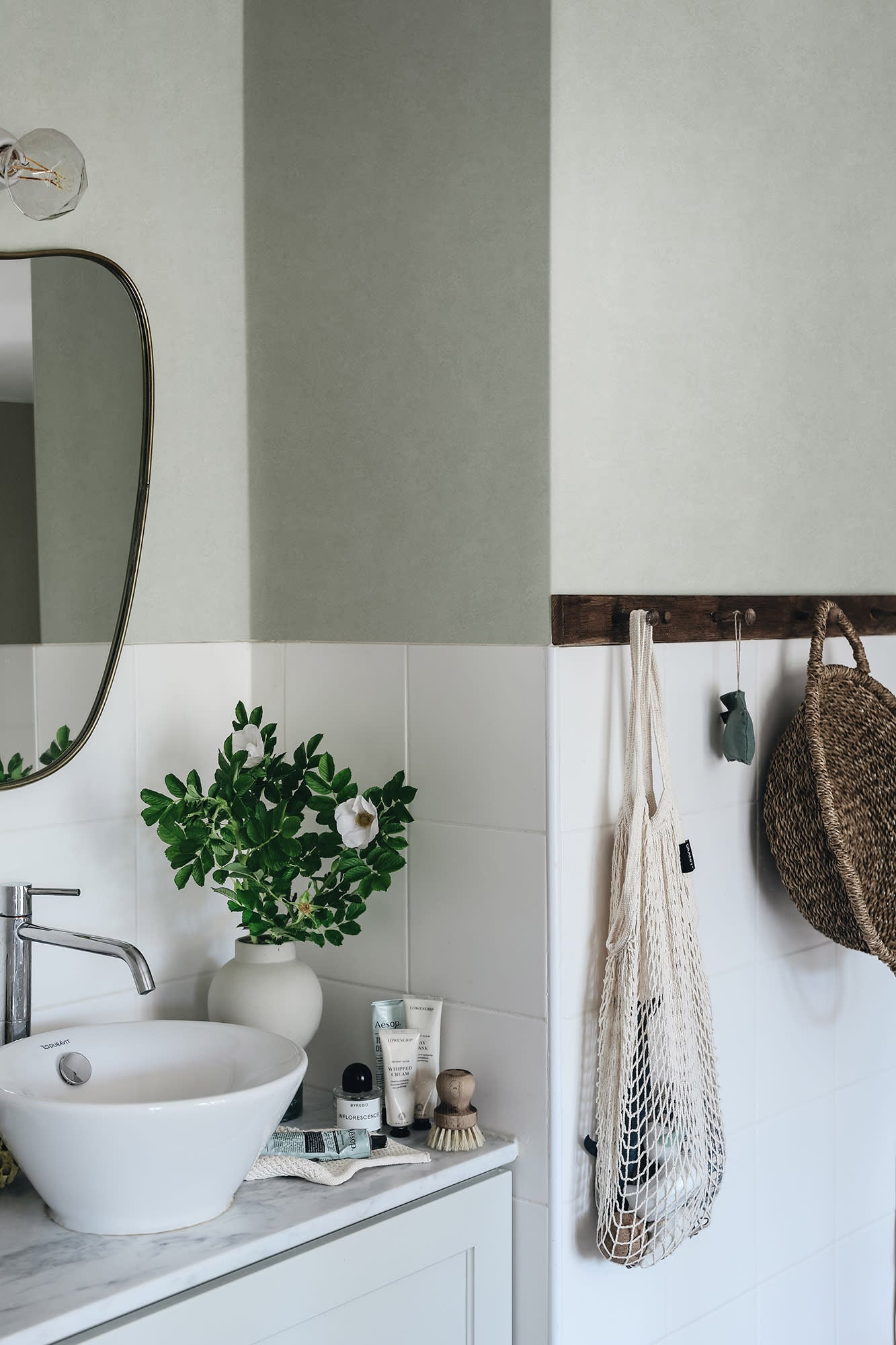 Kalk wallpaper by Sandberg in sage green on a half-tiled bathroom wall above a basin with greenery and a brass mirror