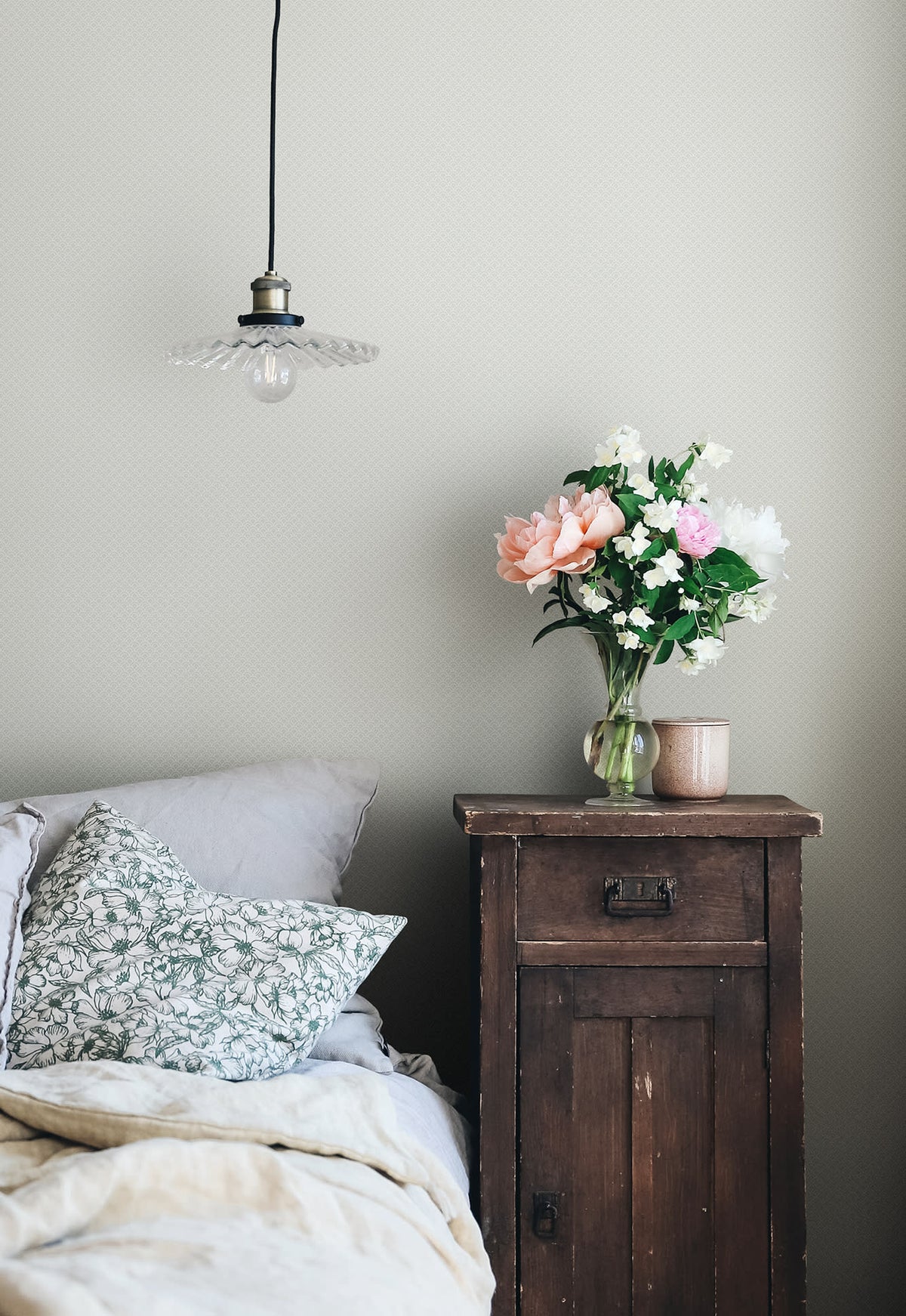 Valv wallpaper by Sandberg in cream in a bedroom beside a rustic timber nightstand with flowers and a glass pendant