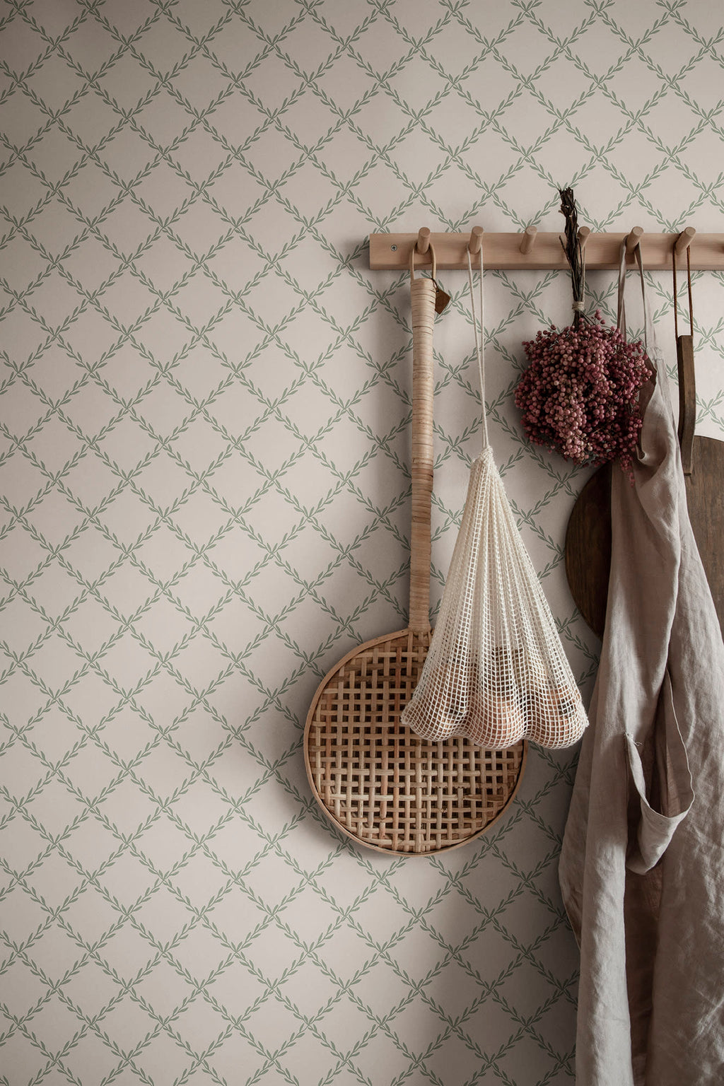 Trellis wallpaper by Sandberg in sage green behind a peg rail holding a linen apron, rattan tray and dried flowers