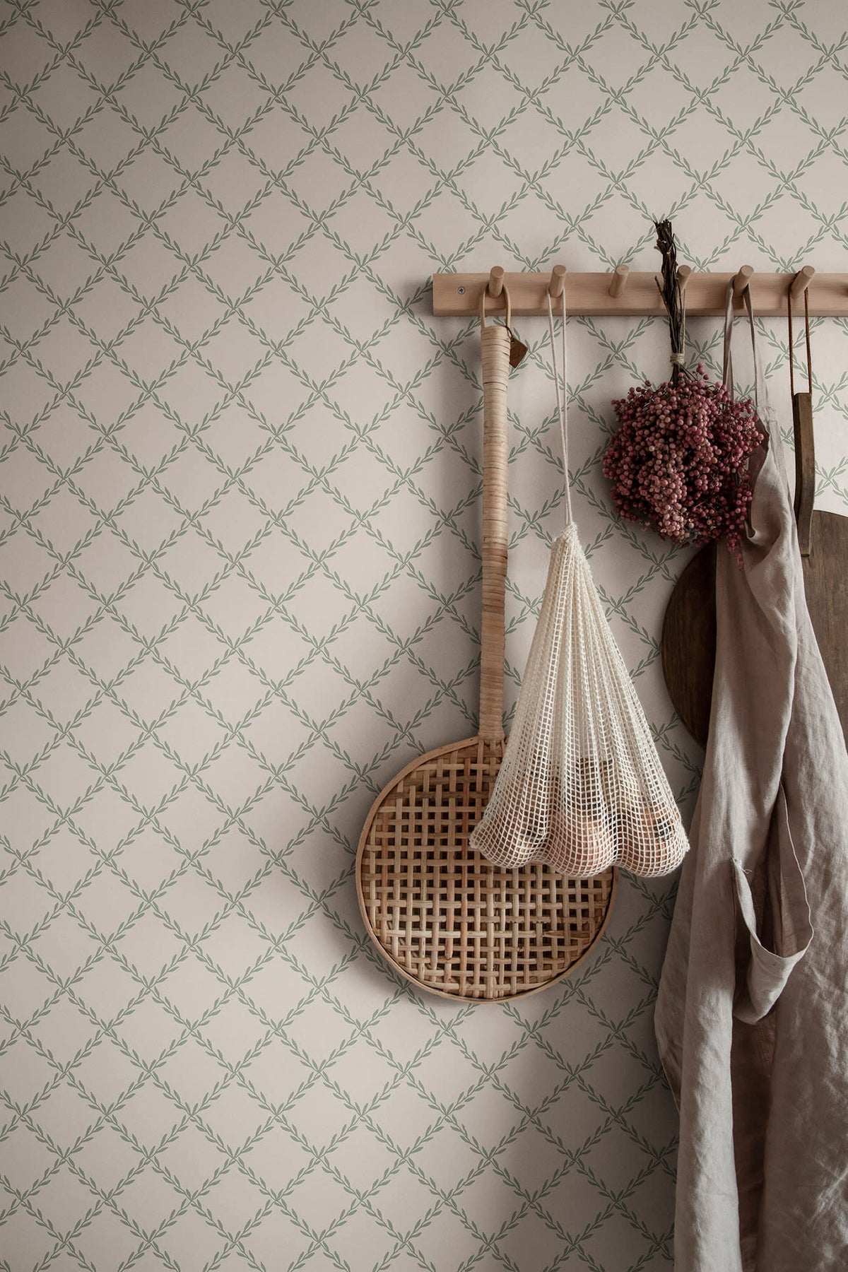 Trellis wallpaper by Sandberg in sage green behind a peg rail holding a linen apron, rattan tray and dried flowers