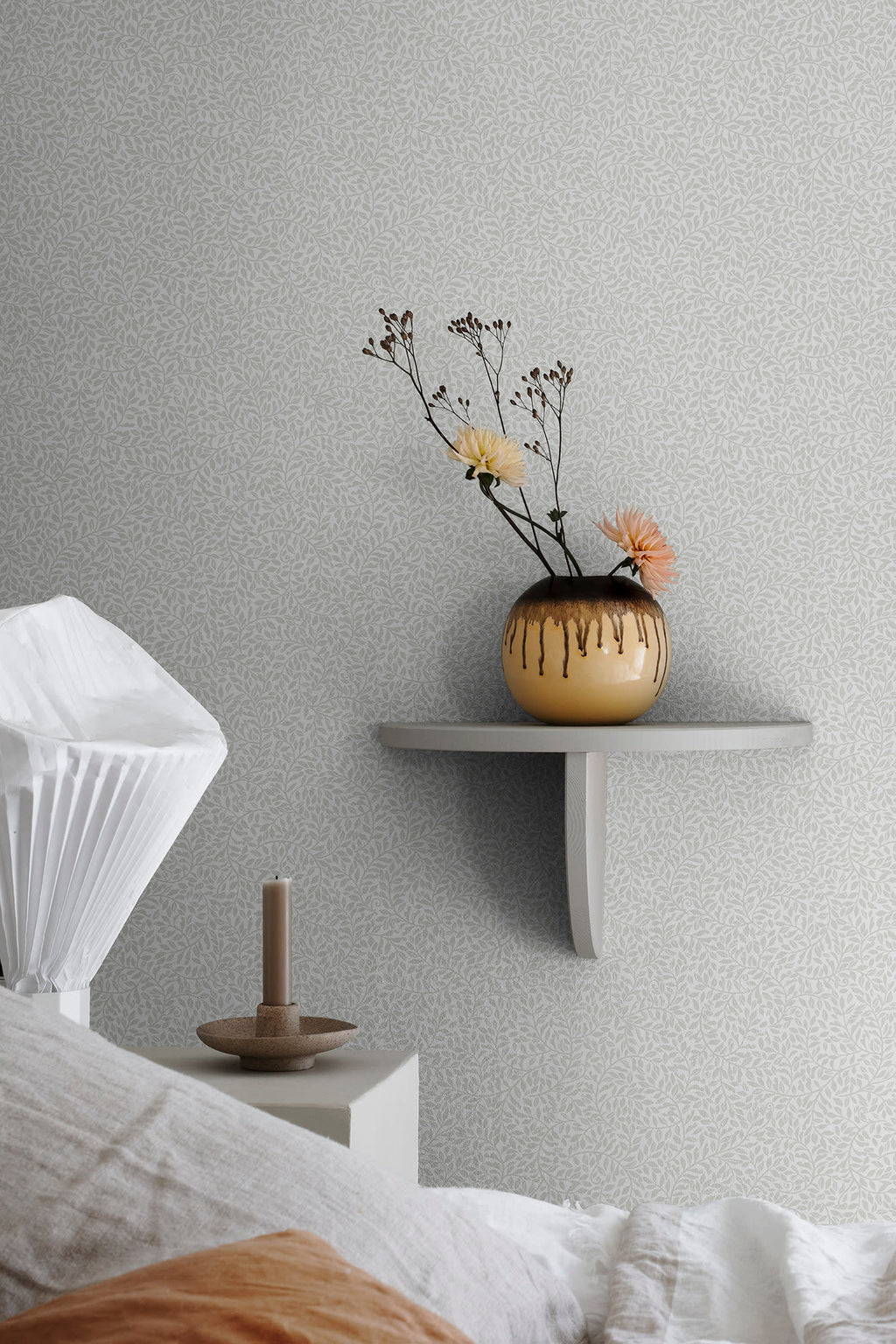 Bladverk wallpaper by Sandberg in grey-green behind a bed, with a floating shelf holding a glazed vase of dried flowers