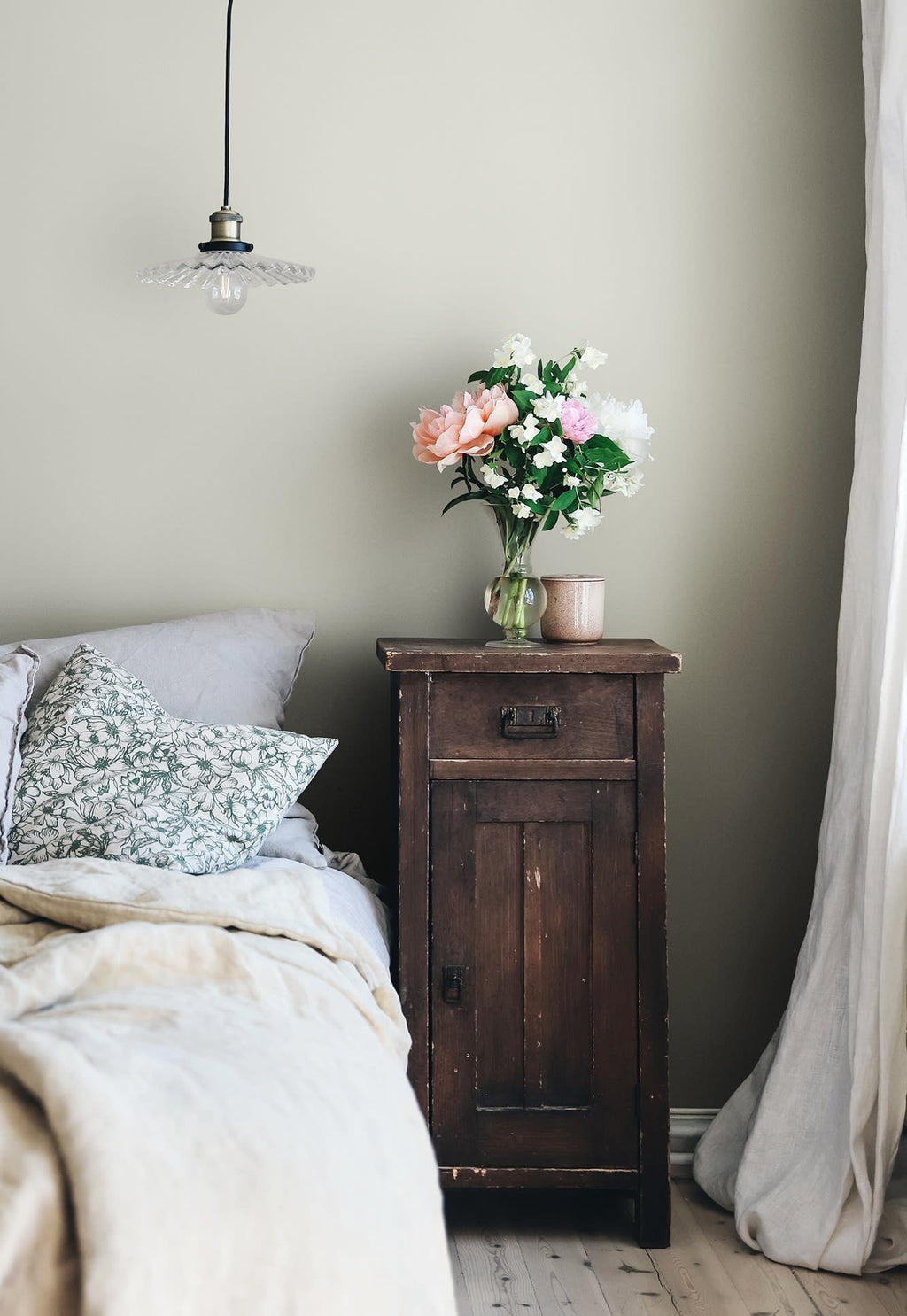 Sten pine branch wallpaper by Sandberg in sage green styled in a bedroom corner with a rustic bedside table and flowers
