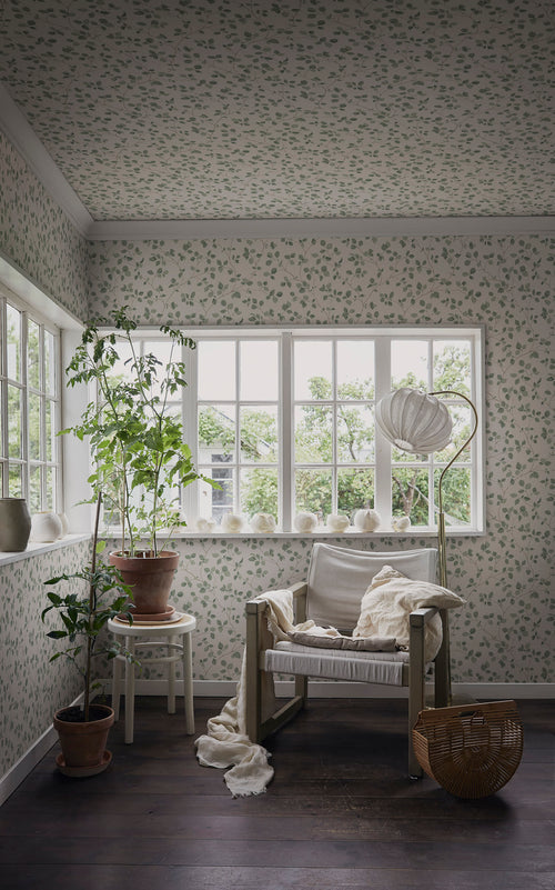Bokskog wallpaper by Sandberg styled in a sunroom, delicate green leaf trails covering walls and ceiling above a linen armchair and potted plants