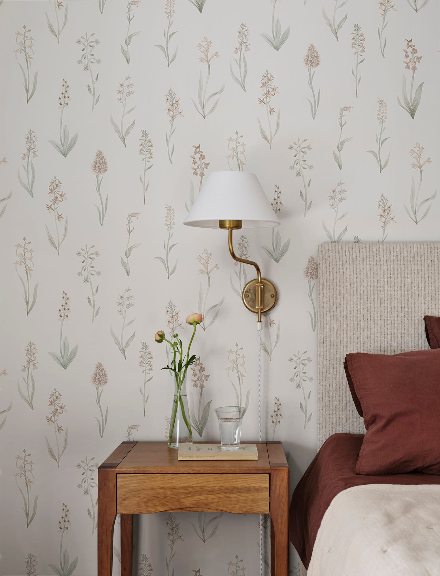 Alma wallpaper by Sandberg, blush botanical stems behind a bed with a brass wall lamp and wooden nightstand