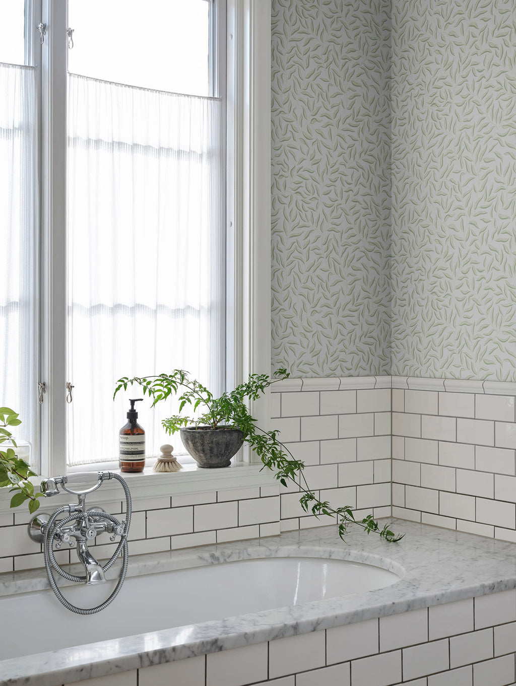 Soft grey-green Karolina leaf wallpaper by Sandberg above white subway tiles by a bathtub and window