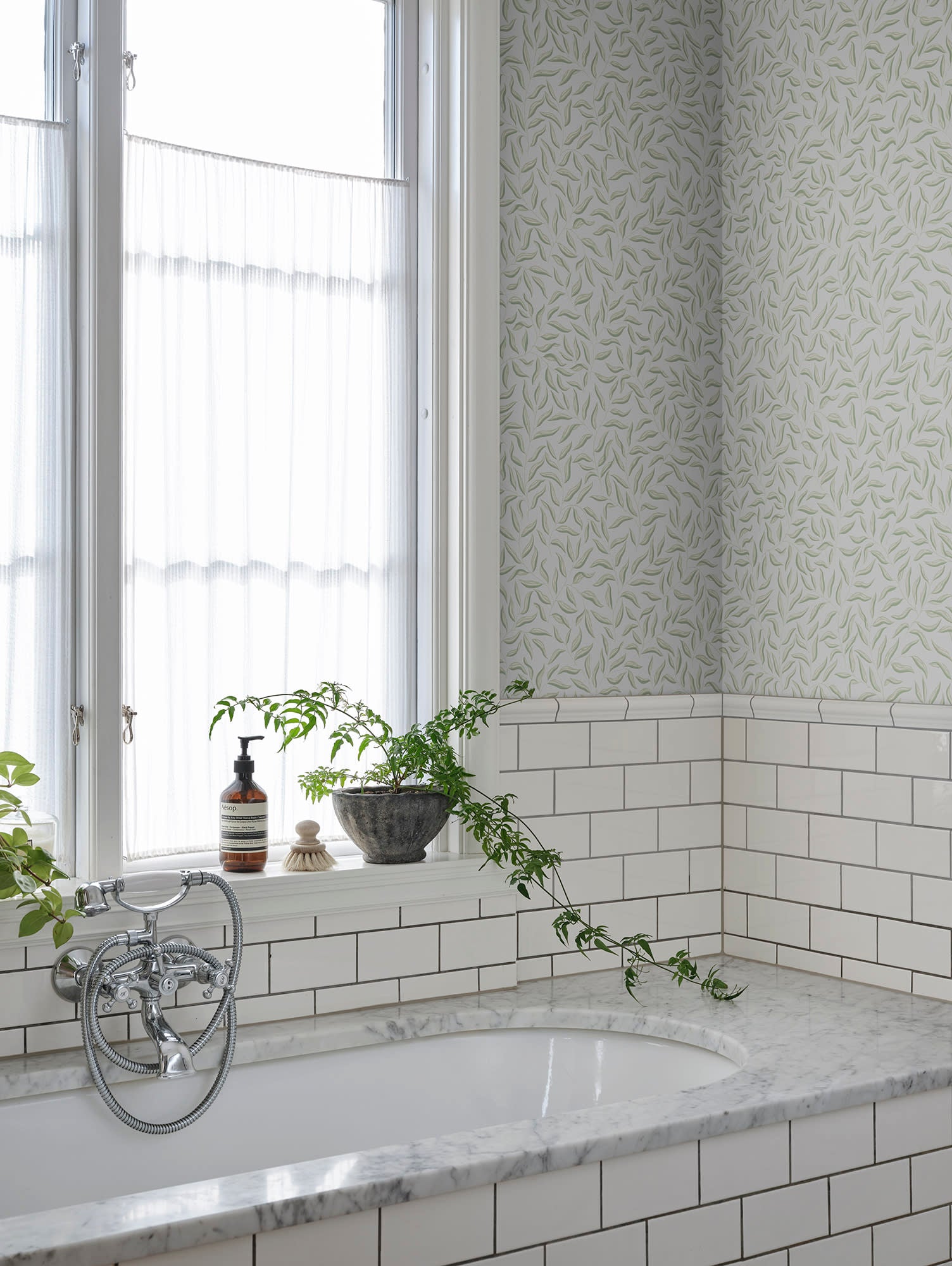 Soft grey-green Karolina leaf wallpaper by Sandberg above white subway tiles by a bathtub and window