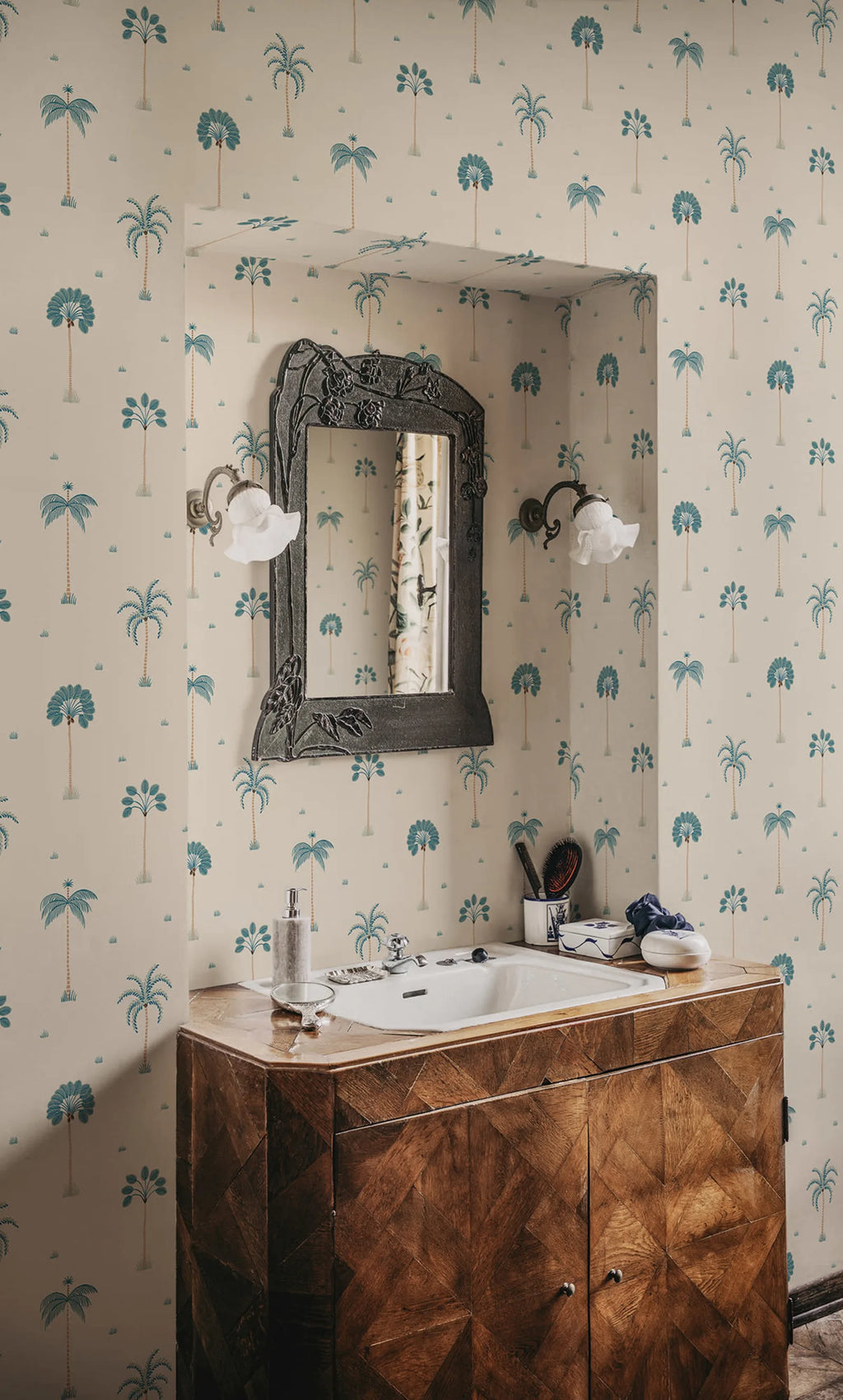 Milo Palms wallpaper by Sandberg in teal on cream, styled in a bathroom around a carved mirror and twin glass sconces