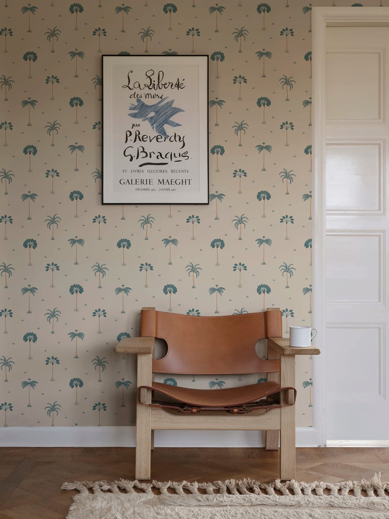 Milo Palms wallpaper by Sandberg in teal on cream, styled behind a tan leather safari chair and framed art print