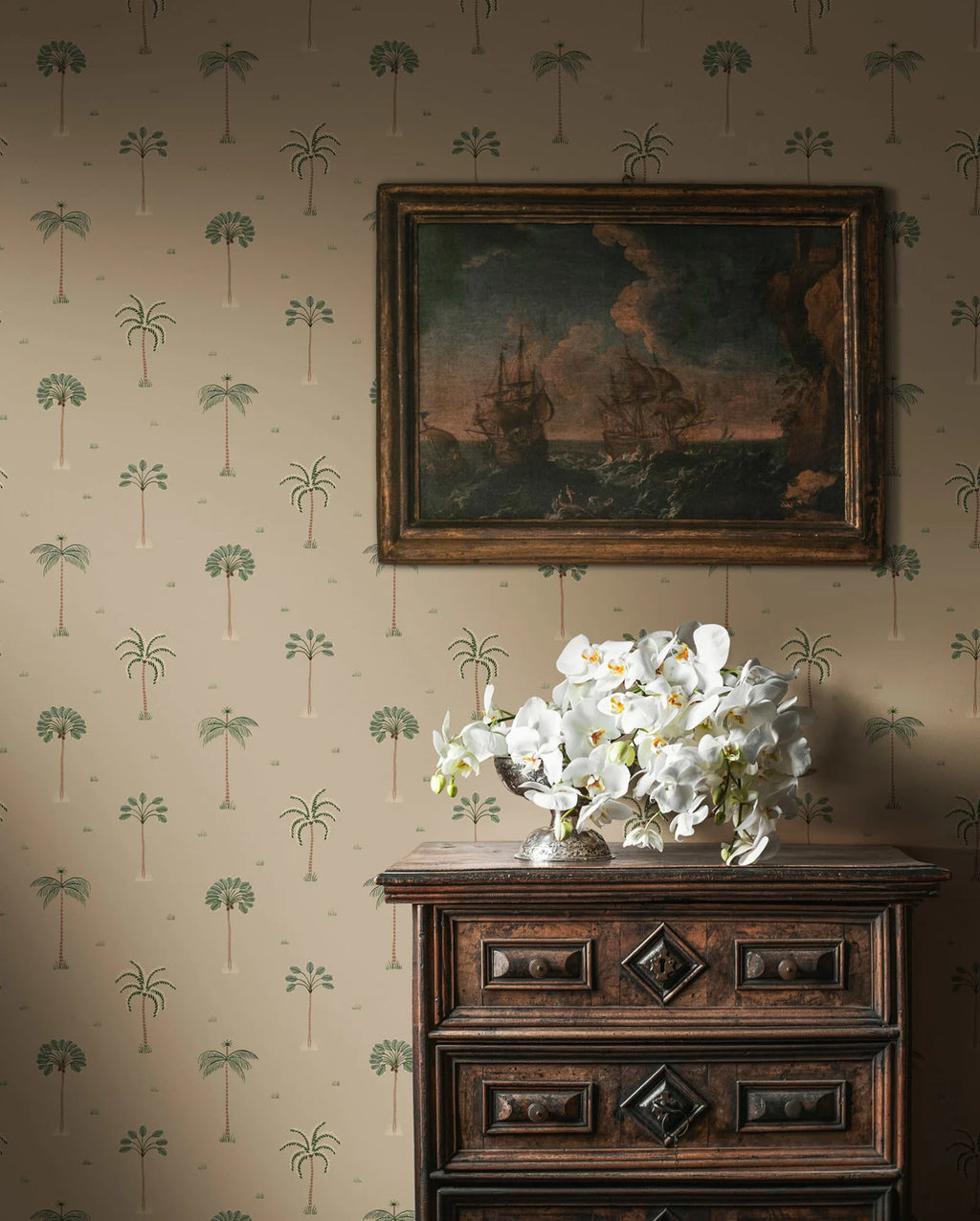 Milo Palms wallpaper by Sandberg in green on sand, styled behind an antique chest topped with a framed seascape and white orchids