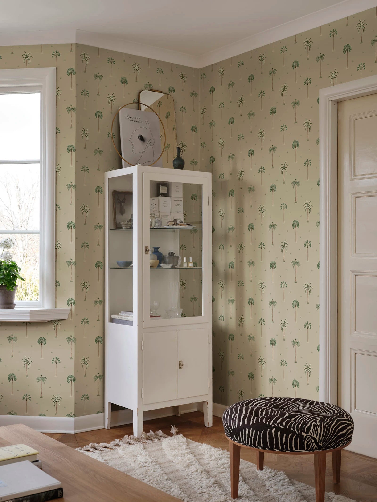 Milo Palms wallpaper by Sandberg in green on sand, styled in a room corner with a white glass display cabinet and zebra stool