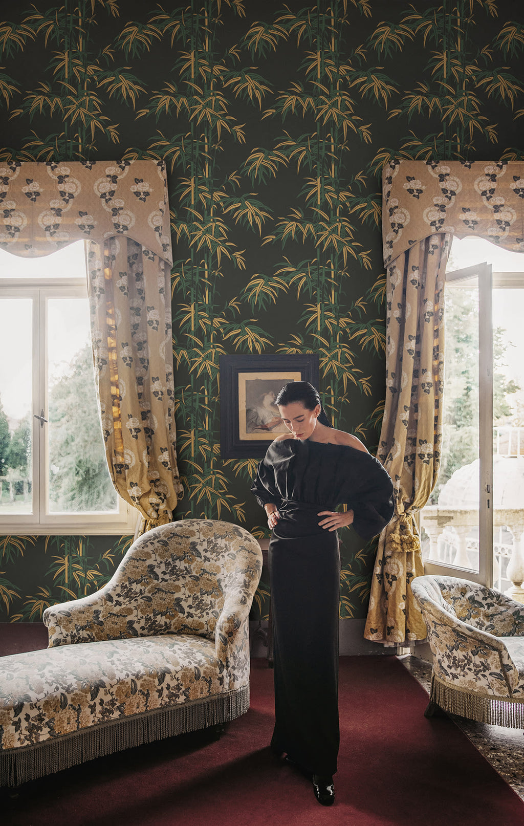 Leon bamboo wallpaper by Sandberg in dark green across a bedroom wall, woman in black between draped windows with floral armchairs