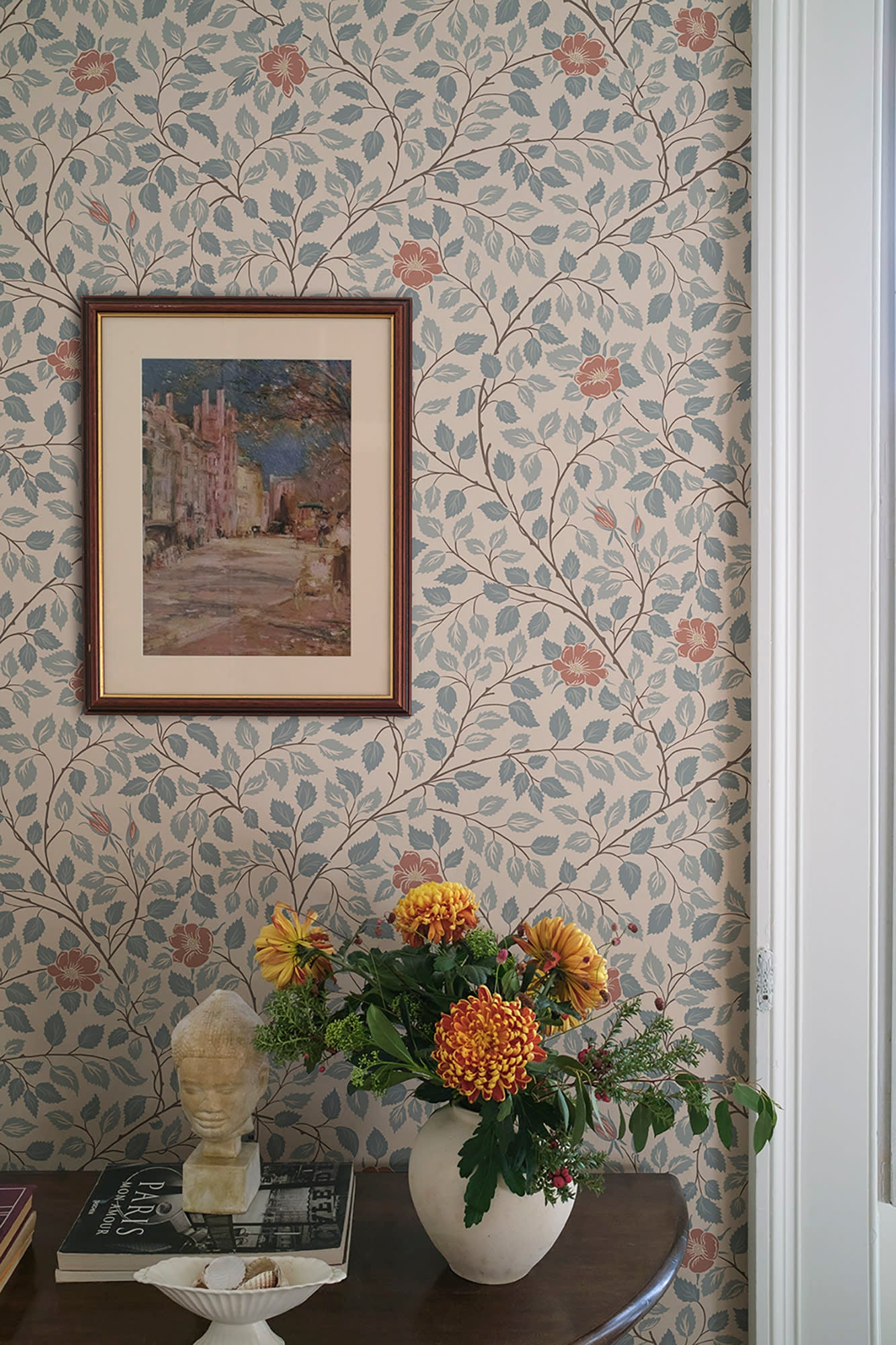 Solliden wallpaper by Sandberg behind a framed print and flowers, blue-leaf and red-rose vines on cream in a styled corner