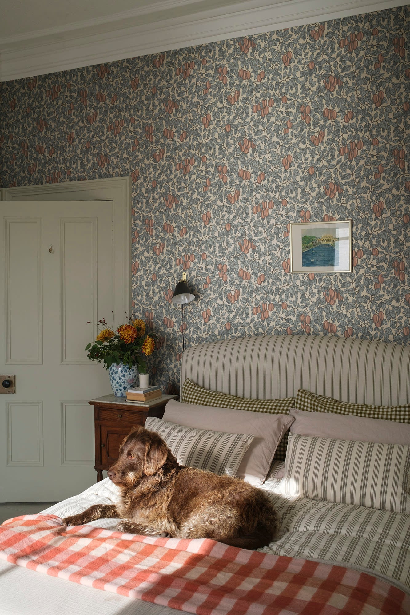 Emrik wallpaper by Sandberg in blue and terracotta in a bedroom with a striped headboard and dog resting on the bed