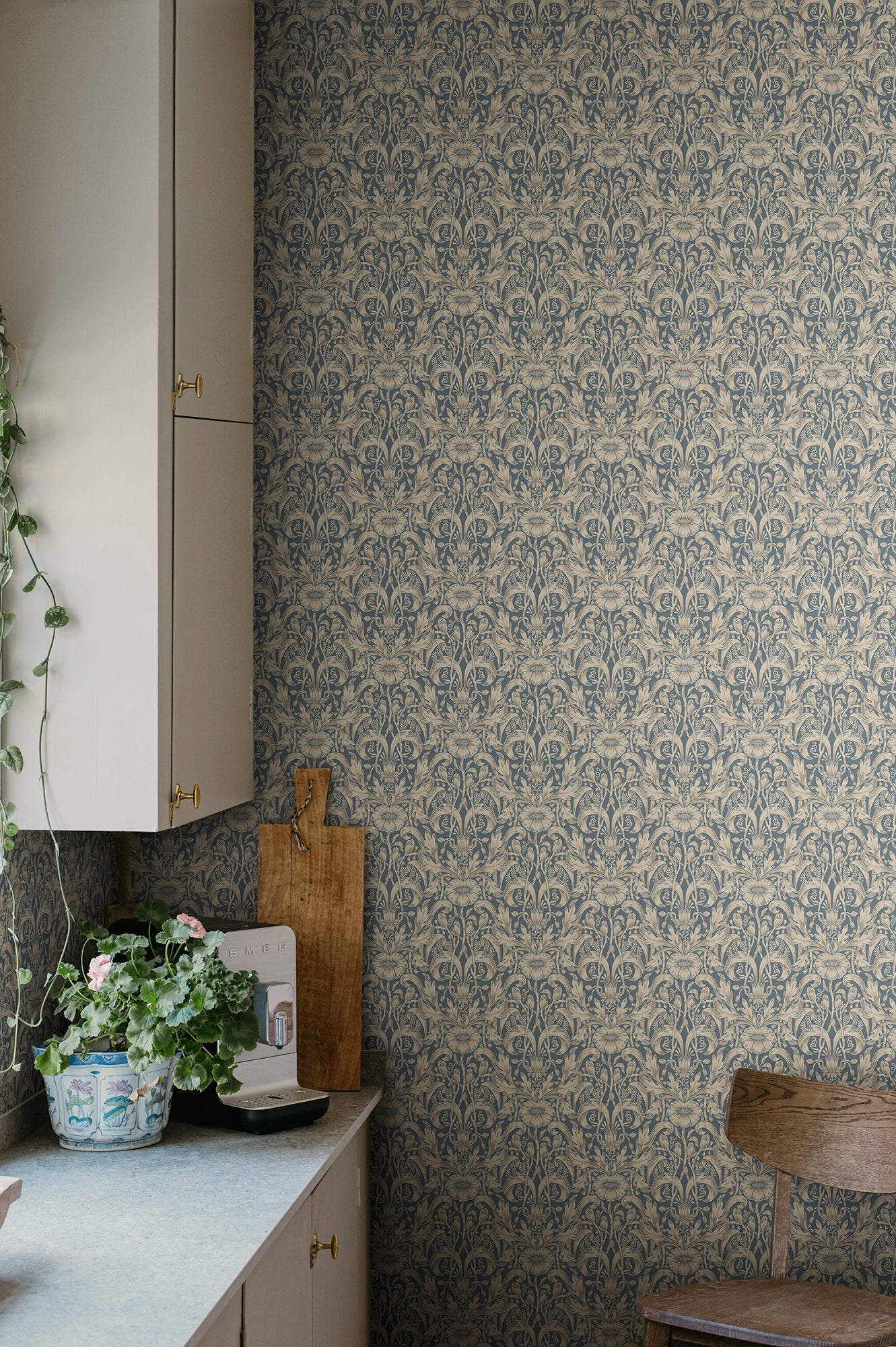 Emil wallpaper by Sandberg in slate blue in a kitchen corner with cream cabinetry and a potted geranium
