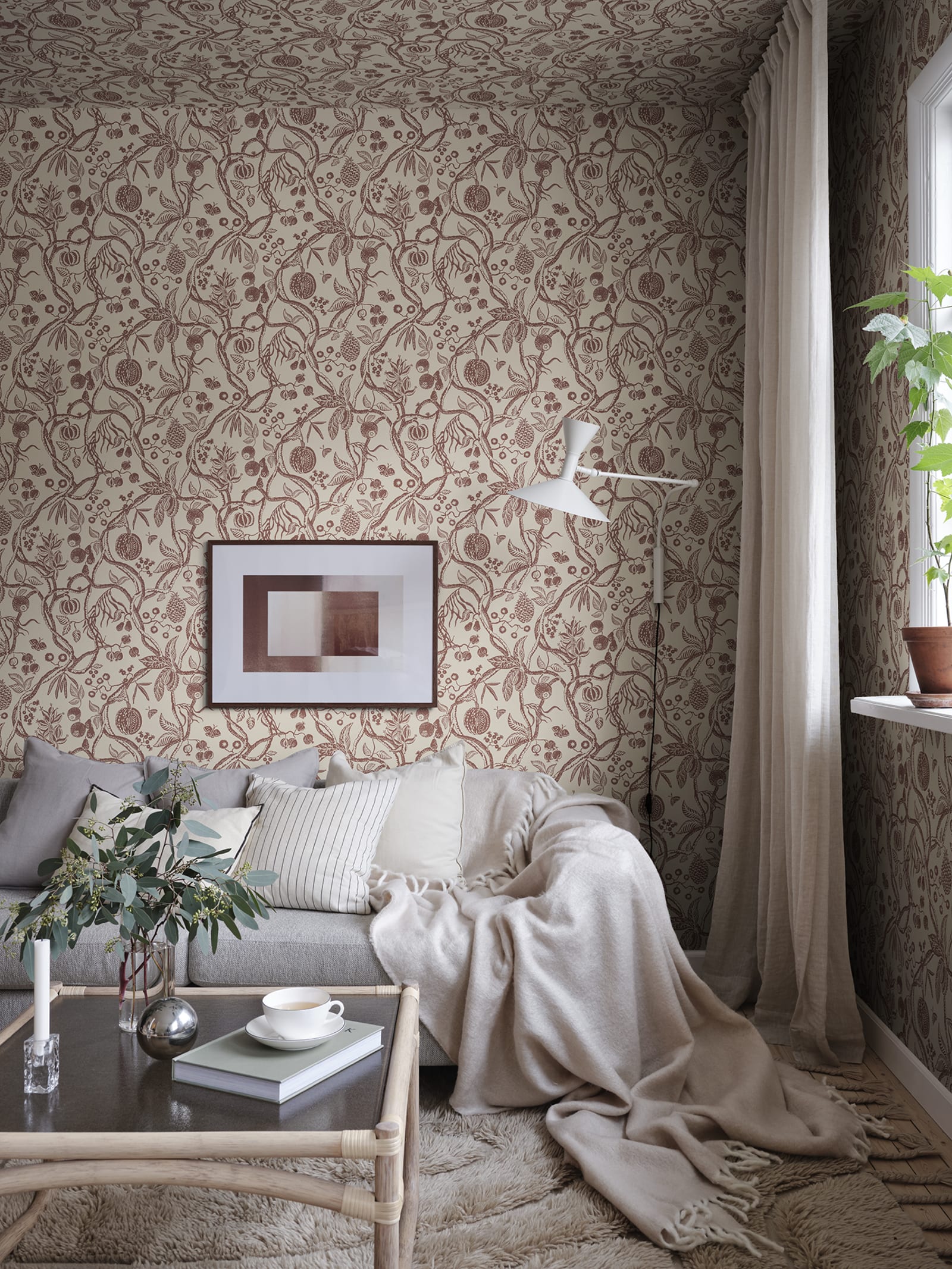 Tree of Life wallpaper by Sandberg in rust-red, styled in a cosy living room behind a grey sofa with framed art and throw