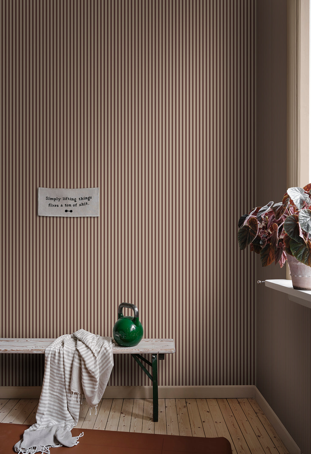 Estelle wallpaper by Sandberg in a brown stripe, styled in a mudroom above a timber bench with a kettlebell and trailing plant