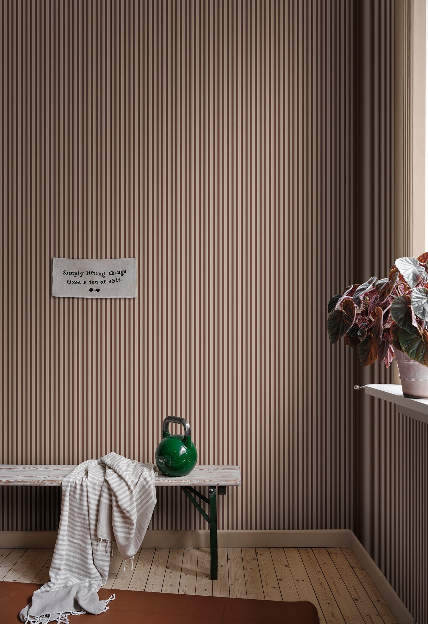 Estelle wallpaper by Sandberg in a brown stripe, styled in a mudroom above a timber bench with a kettlebell and trailing plant