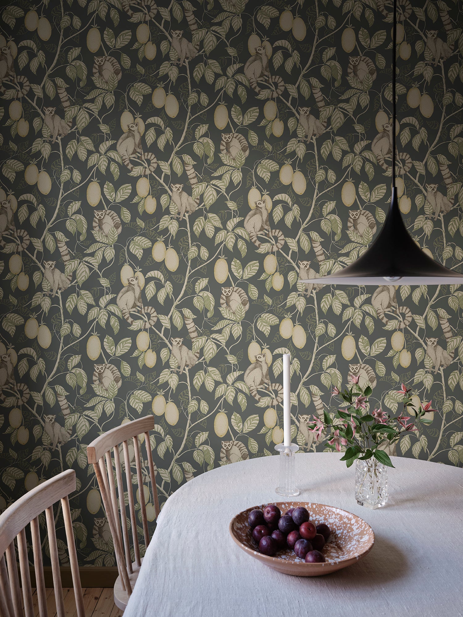 Elliot lemur wallpaper by Sandberg in dark grey-green behind a dining table, pendant lamp over a linen-clad table with plums and flowers