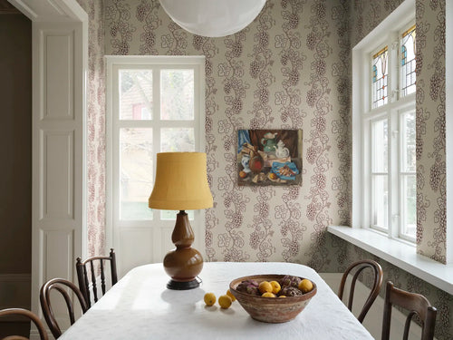 Carla wallpaper by Sandberg in a dining nook with white table, yellow lamp and stained-glass window, trailing grapevine design