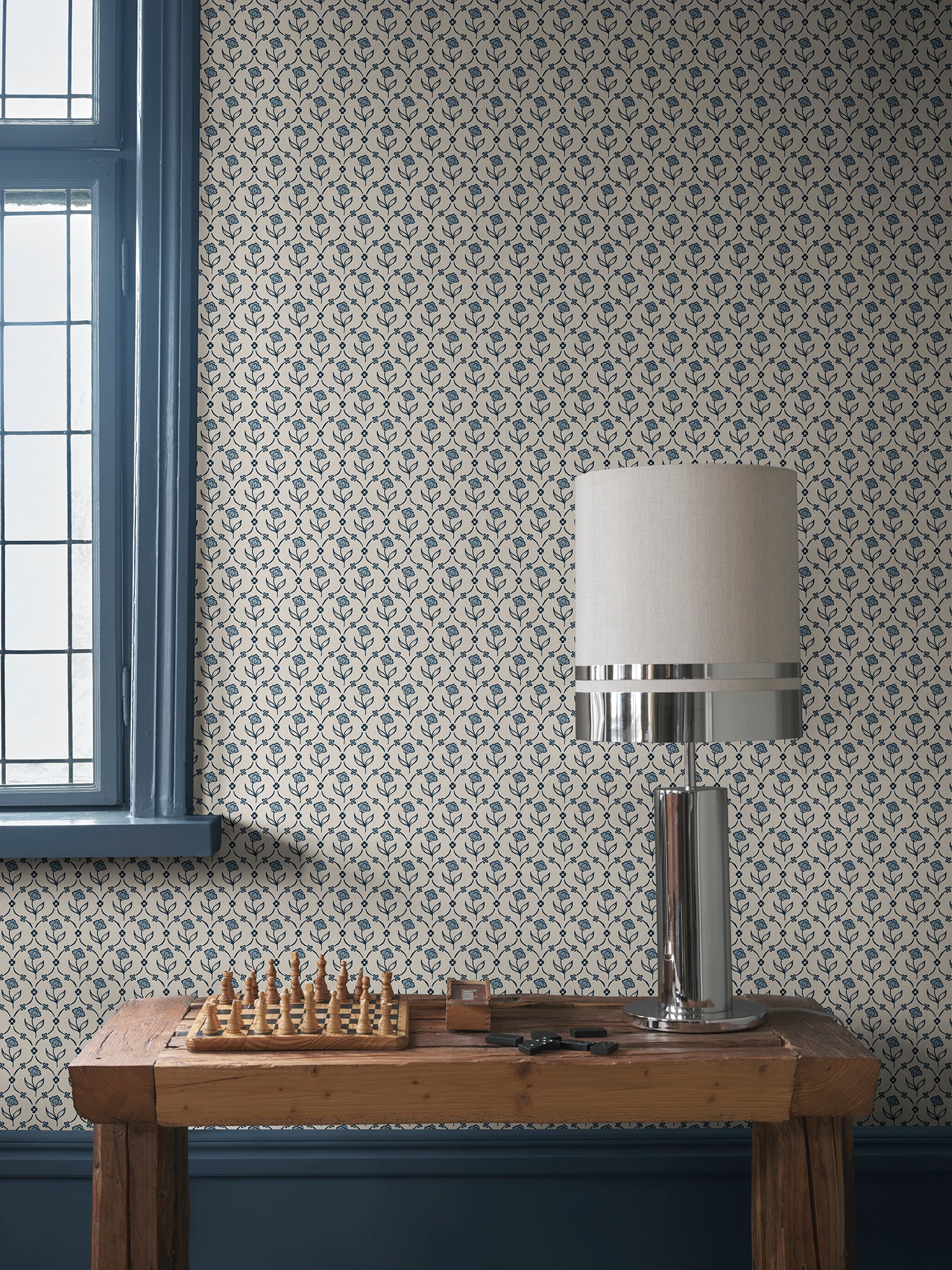 Elena blue floral wallpaper by Sandberg behind a pine console with a chrome lamp and wooden chess set beside a window