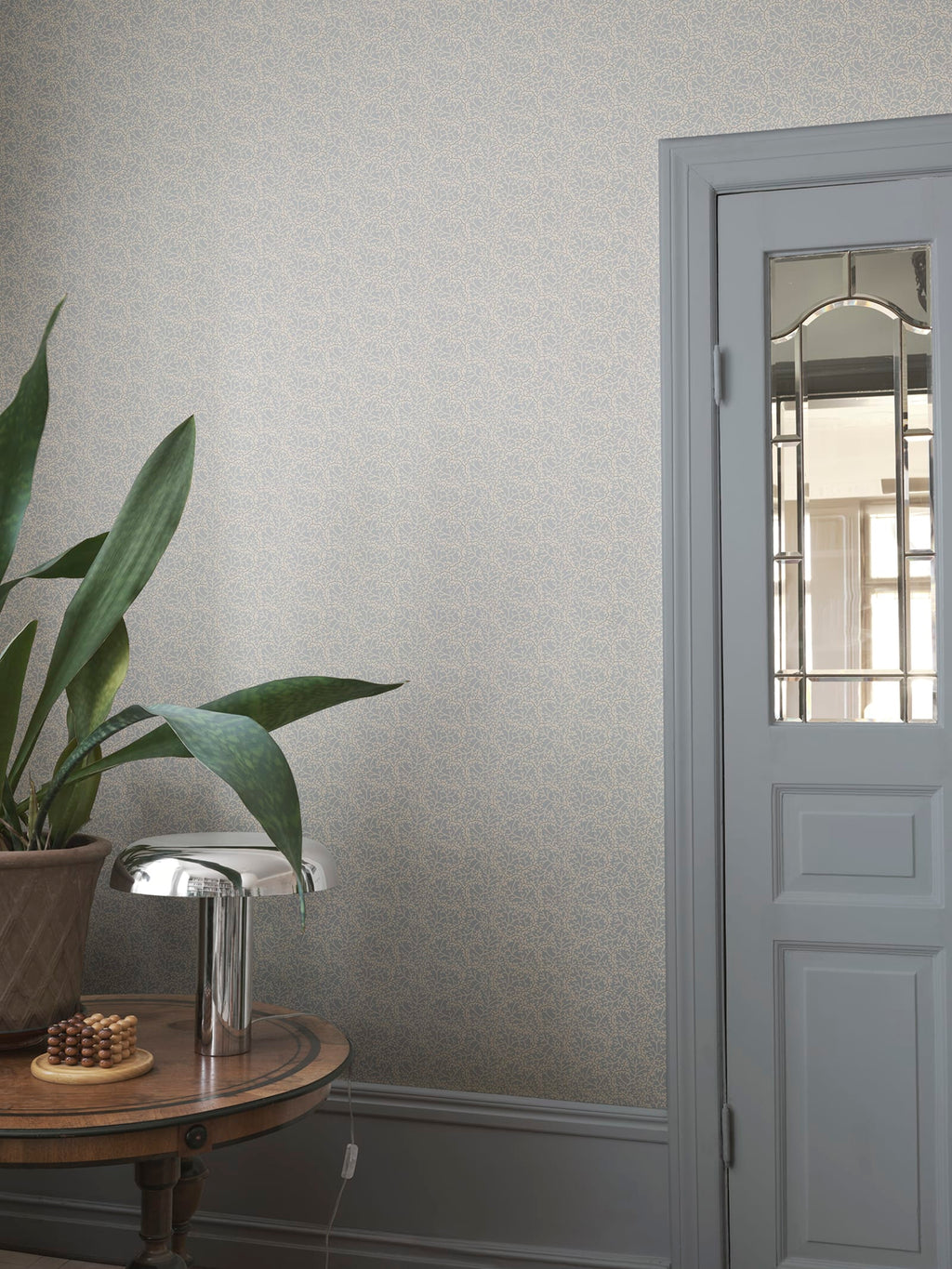 Coral wallpaper by Sandberg in muted grey-blue, styled in an entryway beside a glazed door with snake plant and chrome lamp