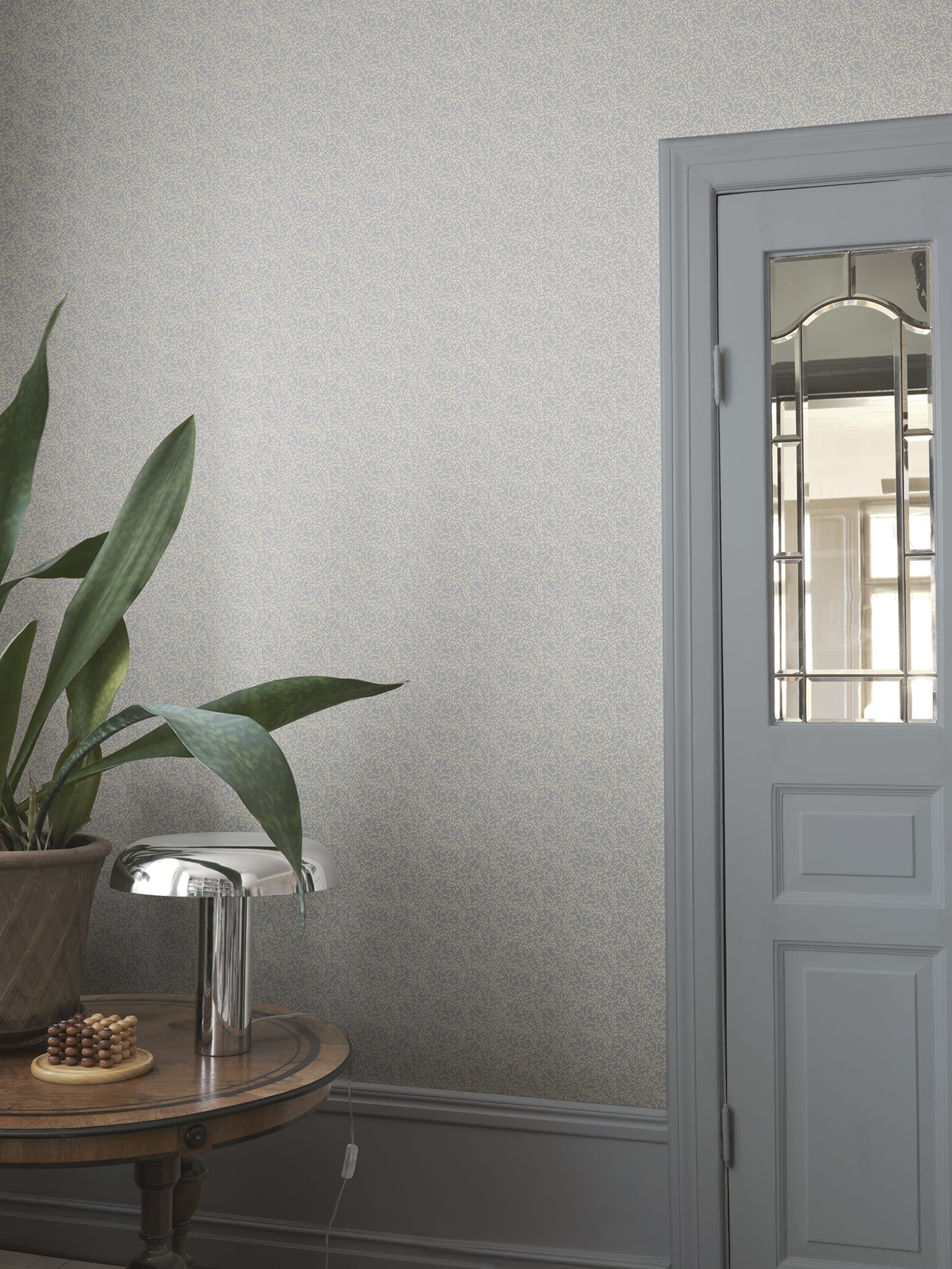 Coral wallpaper by Sandberg in muted grey-blue, styled in an entryway beside a glazed door with snake plant and chrome lamp