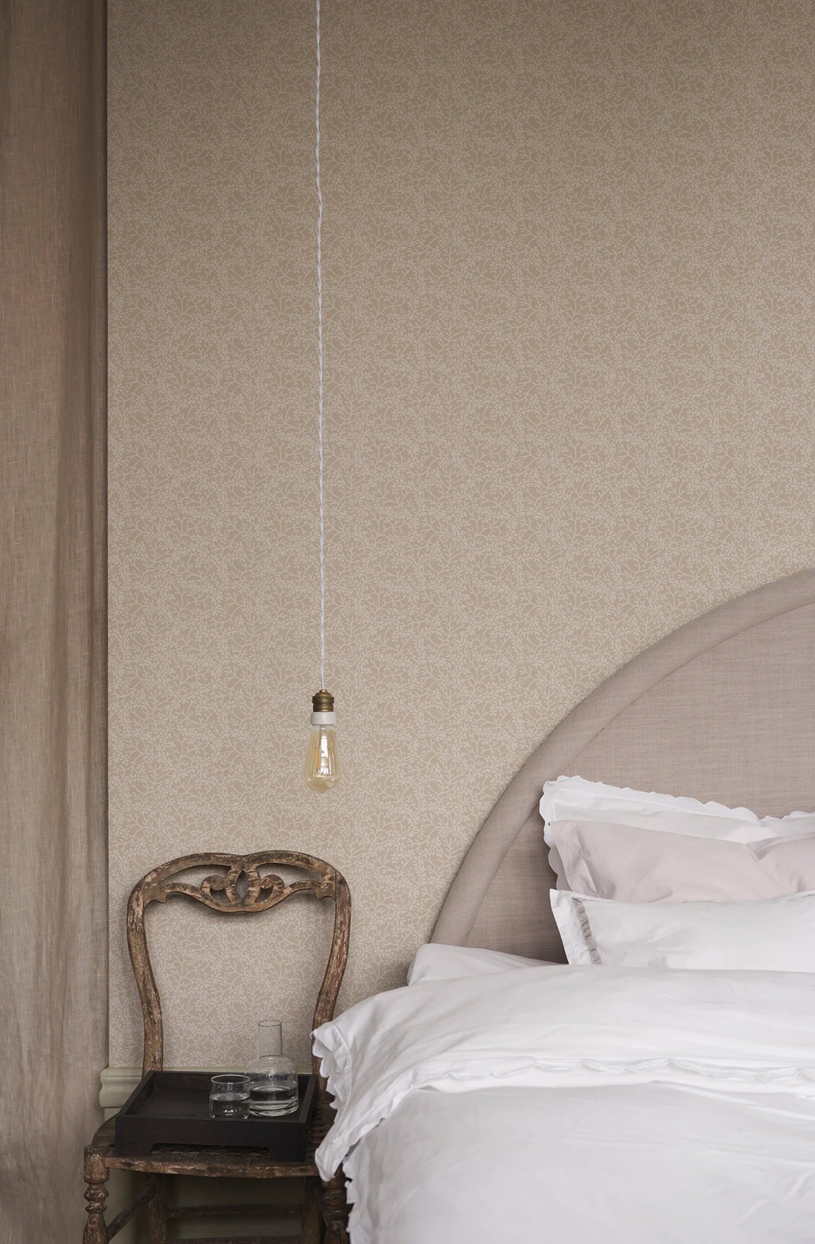 Coral wallpaper by Sandberg in tonal sand, small coral-branch motif styled behind a linen bed with antique chair and pendant bulb