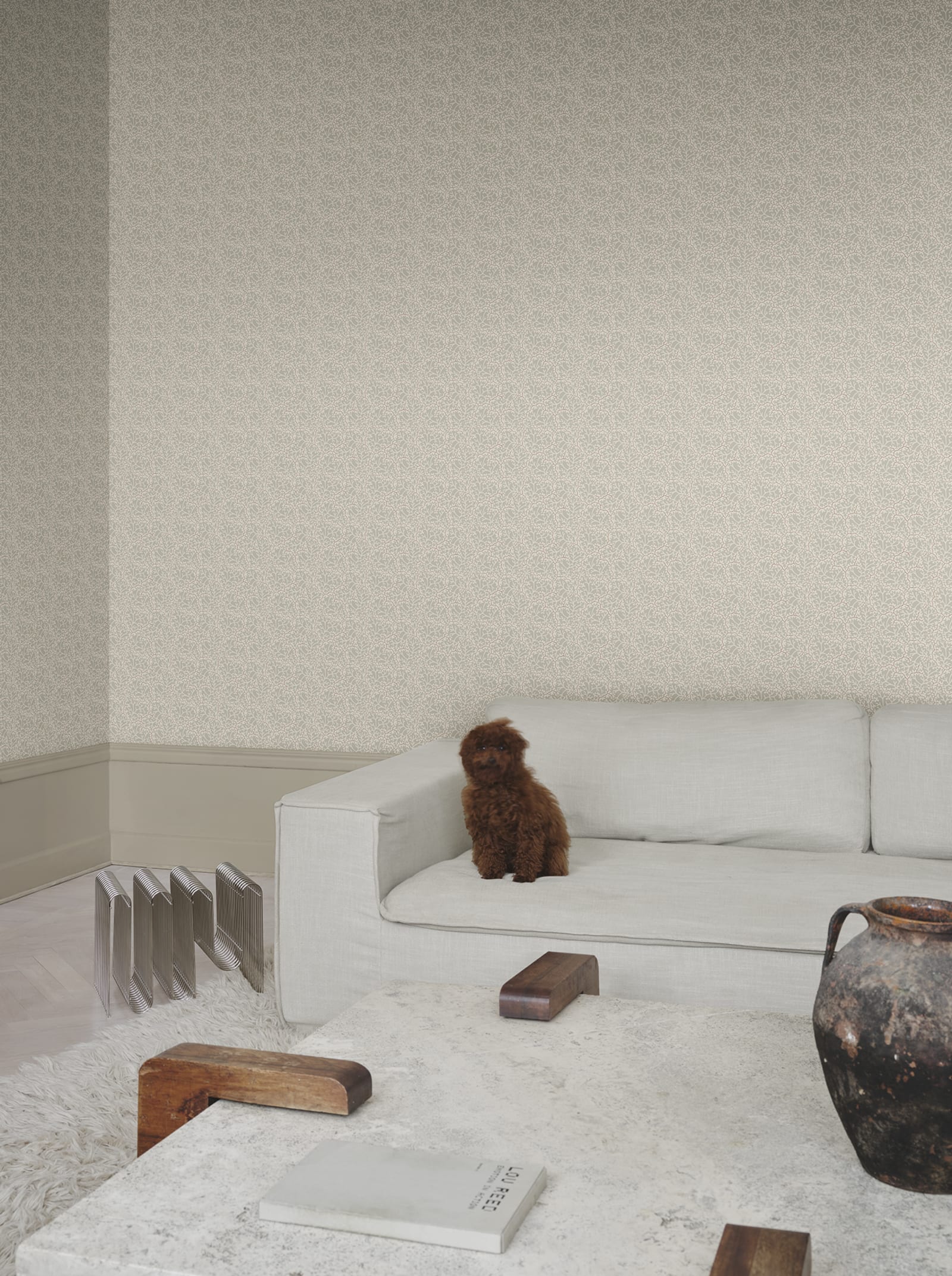 Coral wallpaper by Sandberg in pale sand, styled in a living room above a white sofa with a small brown poodle