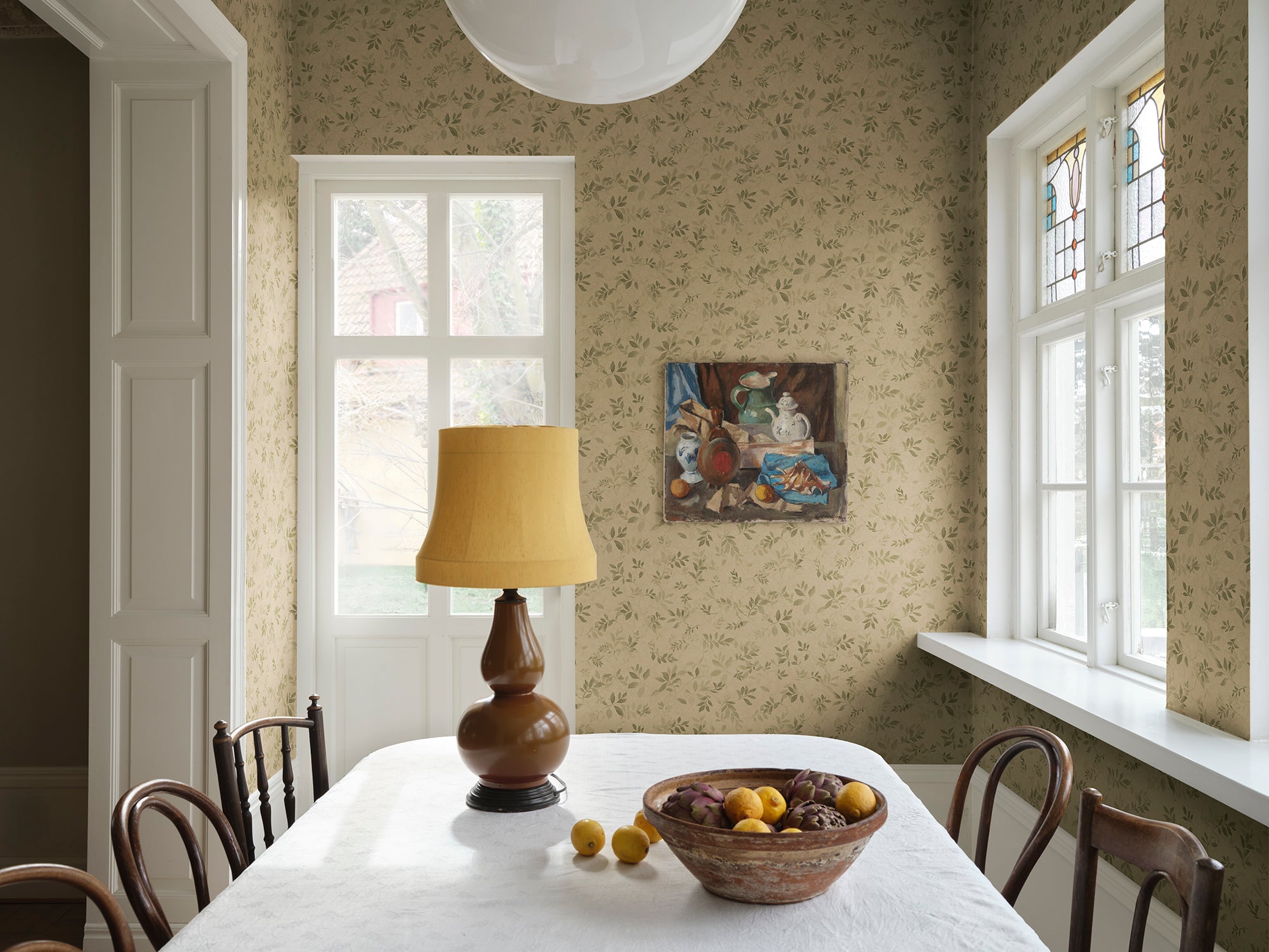 Mila wallpaper by Sandberg in green on sand, styled in a dining room with a white-clothed table, brown lamp and stained-glass window