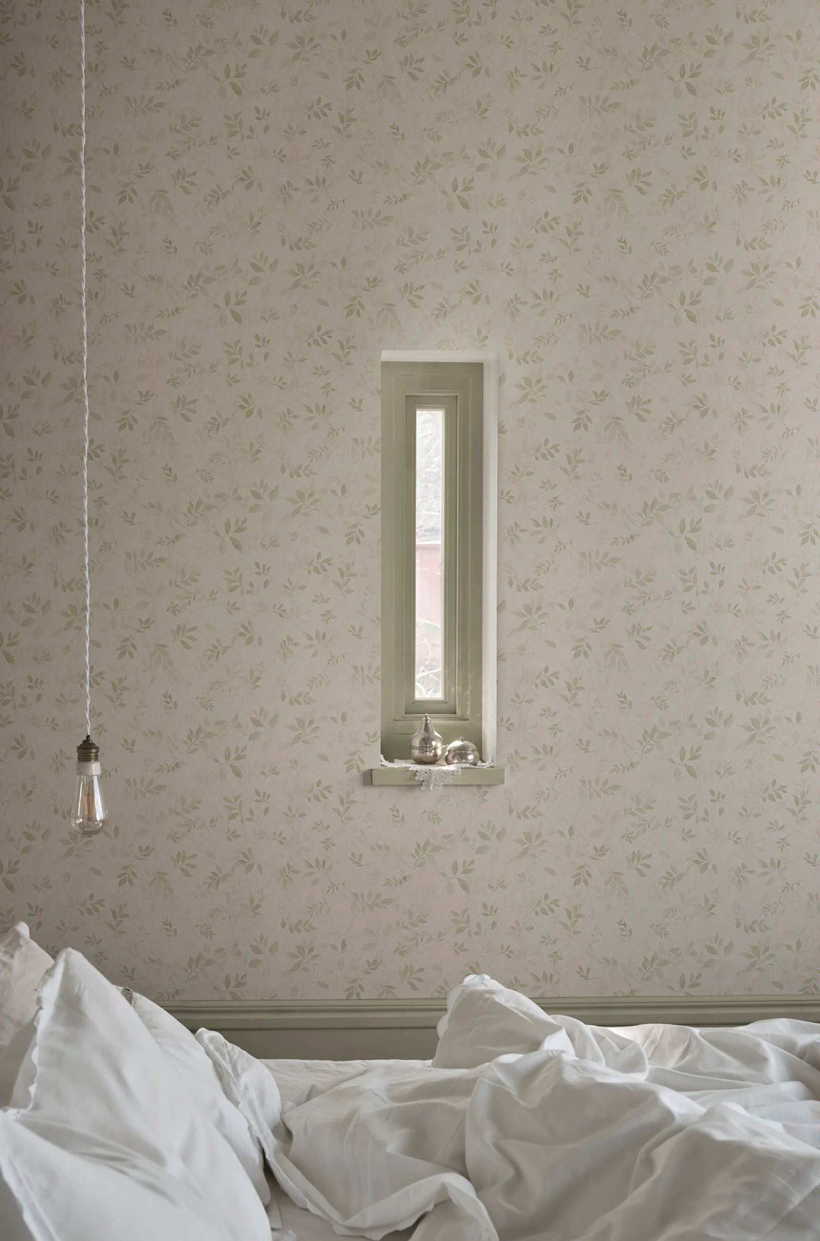 Mila wallpaper by Sandberg in sage on cream, styled in a bedroom with a slim recessed window and rumpled white bedding