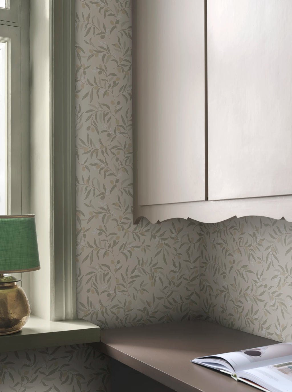 Olivia wallpaper by Sandberg in sage-green olive branches on cream, styled behind a window and kitchen cabinetry with a green lamp