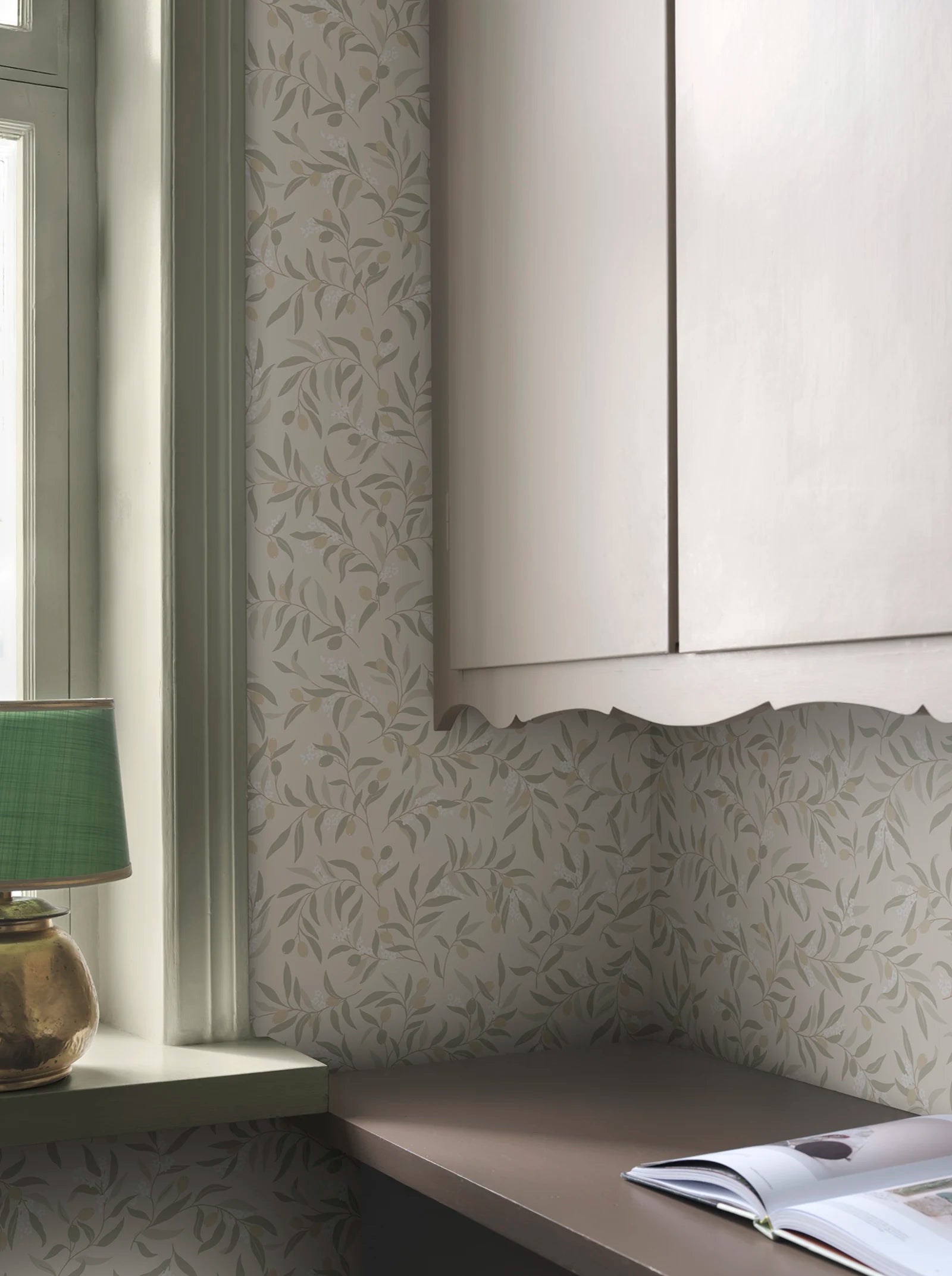 Olivia wallpaper by Sandberg in sage-green olive branches on cream, styled behind a window and kitchen cabinetry with a green lamp