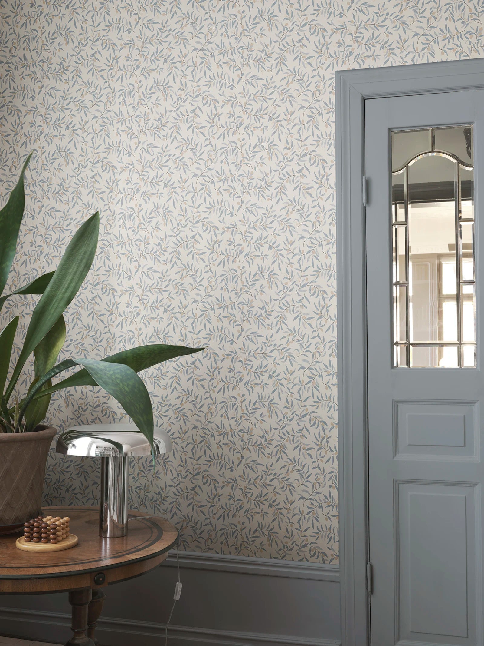 Olivia wallpaper by Sandberg in blue-grey olive branches, styled in an entryway beside a glazed door and snake plant