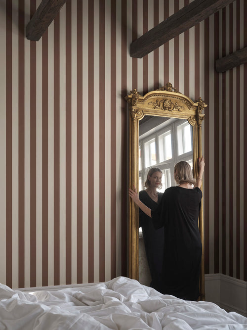 Brown striped Sam wallpaper by Sandberg behind a tall gold-framed mirror in a beamed bedroom