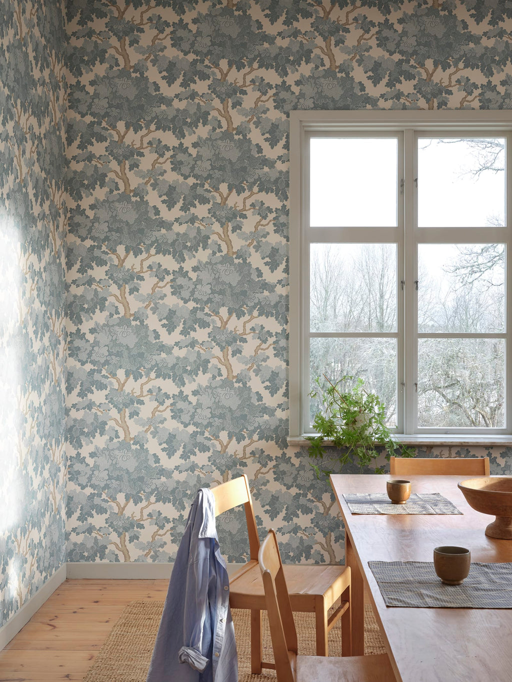 Eken wallpaper by Sandberg, dusty-blue oak trees on cream, styled in a dining room beside a window with timber table and chairs