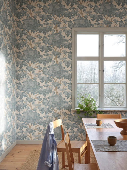 Eken wallpaper by Sandberg, dusty-blue oak trees on cream, styled in a dining room beside a window with timber table and