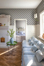 Annie wallpaper by Sandberg in blue above white panelling in a sunny sitting room with a gingham sofa, tiled stove and green side table