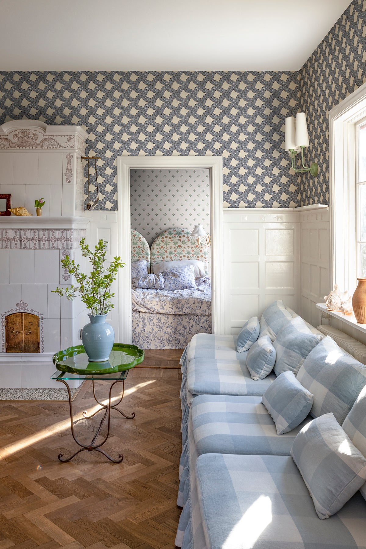 Annie wallpaper by Sandberg in blue above white panelling in a sunny sitting room with a gingham sofa, tiled stove and green side table