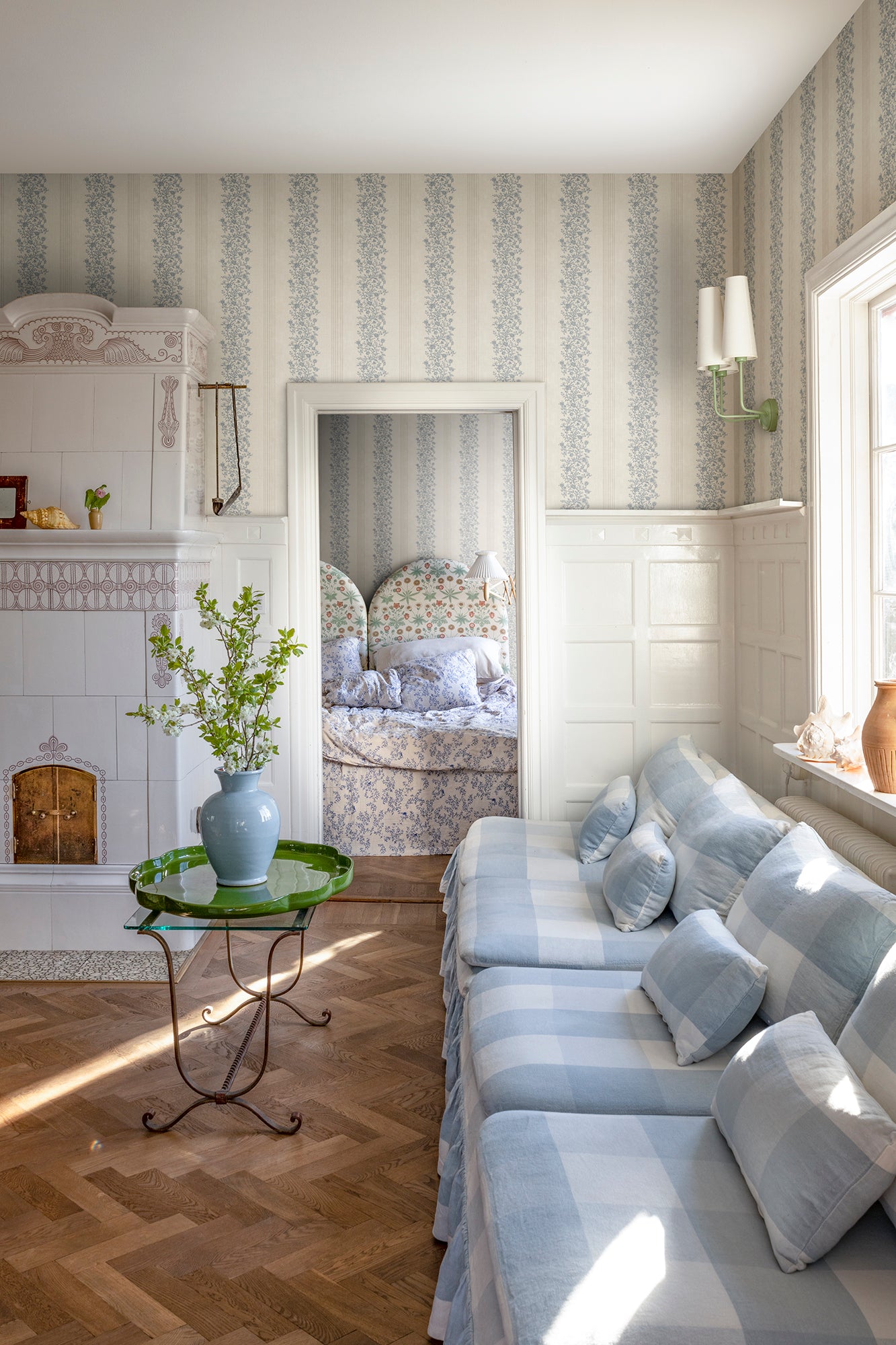 Natalie wallpaper by Sandberg styled in a sunlit living room with check sofa and green side table, blue floral-stripe design