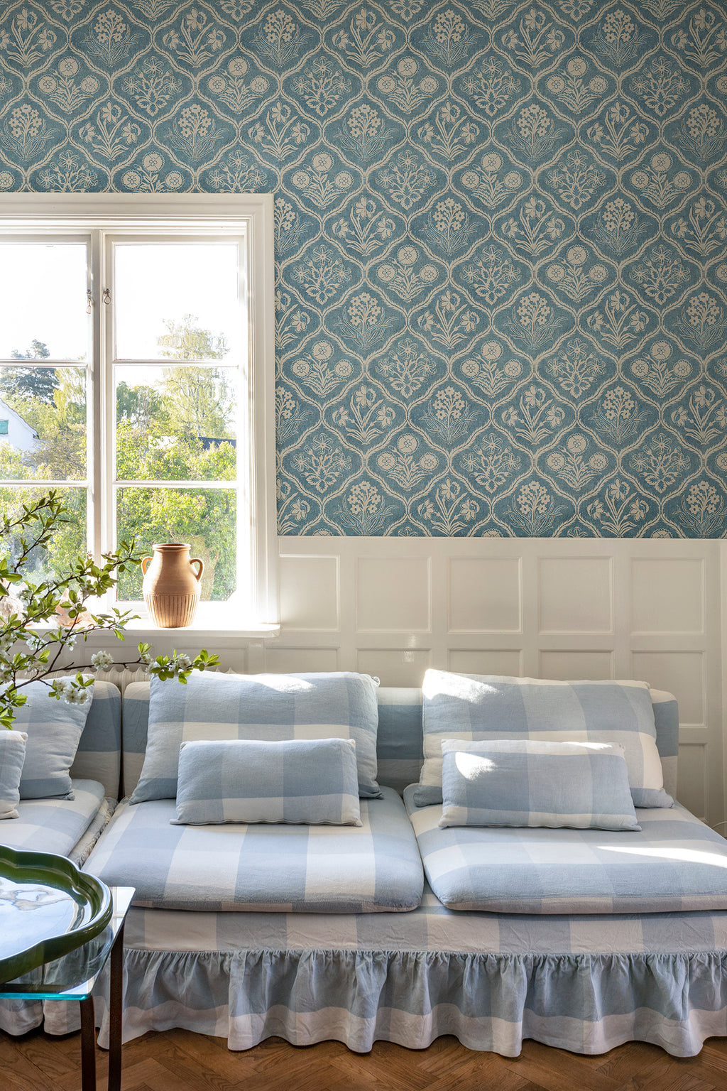 Rosa wallpaper by Sandberg above white panelling with a blue check sofa and window, blue floral-trellis pattern
