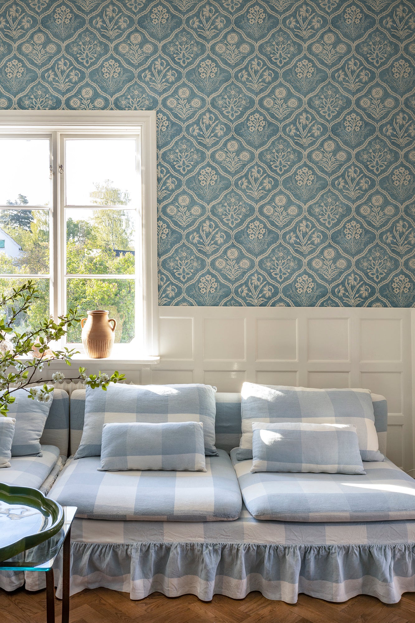 Rosa wallpaper by Sandberg above white panelling with a blue check sofa and window, blue floral-trellis pattern