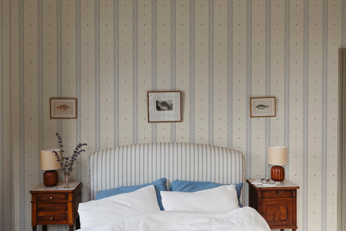 Austine wallpaper by Sandberg in Blue behind a striped headboard in a calm bedroom with framed prints