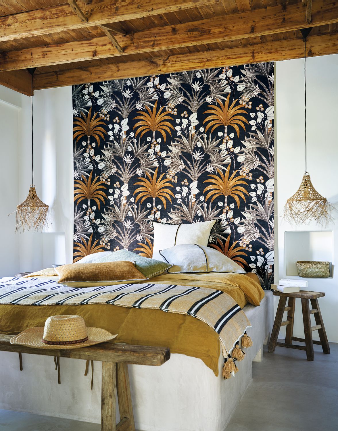 Mirissa wallpaper by Casamance in ink navy and ochre, a palm and blossom print styled behind a bed under timber beams