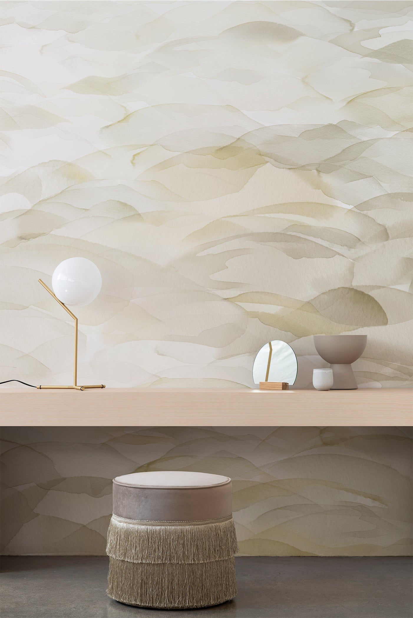 Dune wallpaper by Emma Hayes in fog beige and cream watercolour layers above a timber shelf with brass lamp and a fringed ottoman below